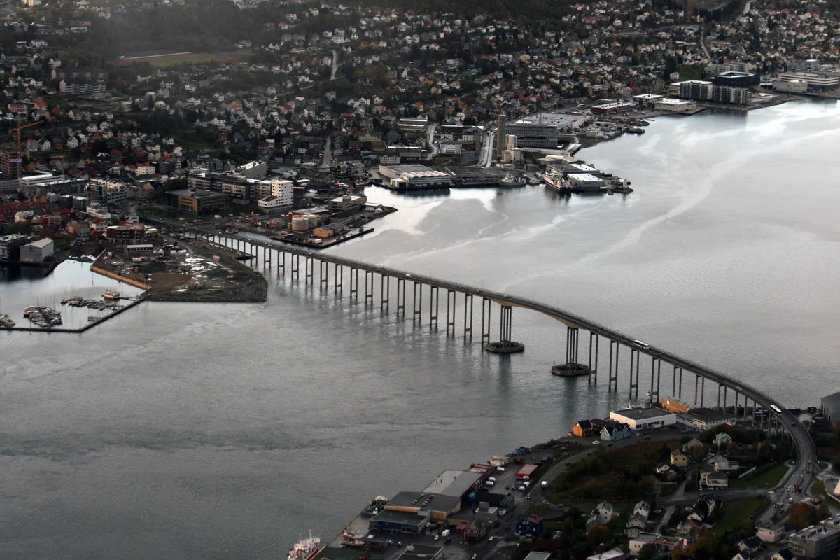 Ponte curva sobre o estreito, com pilares, as duas margens urbanizadas e edifícios junto à água.