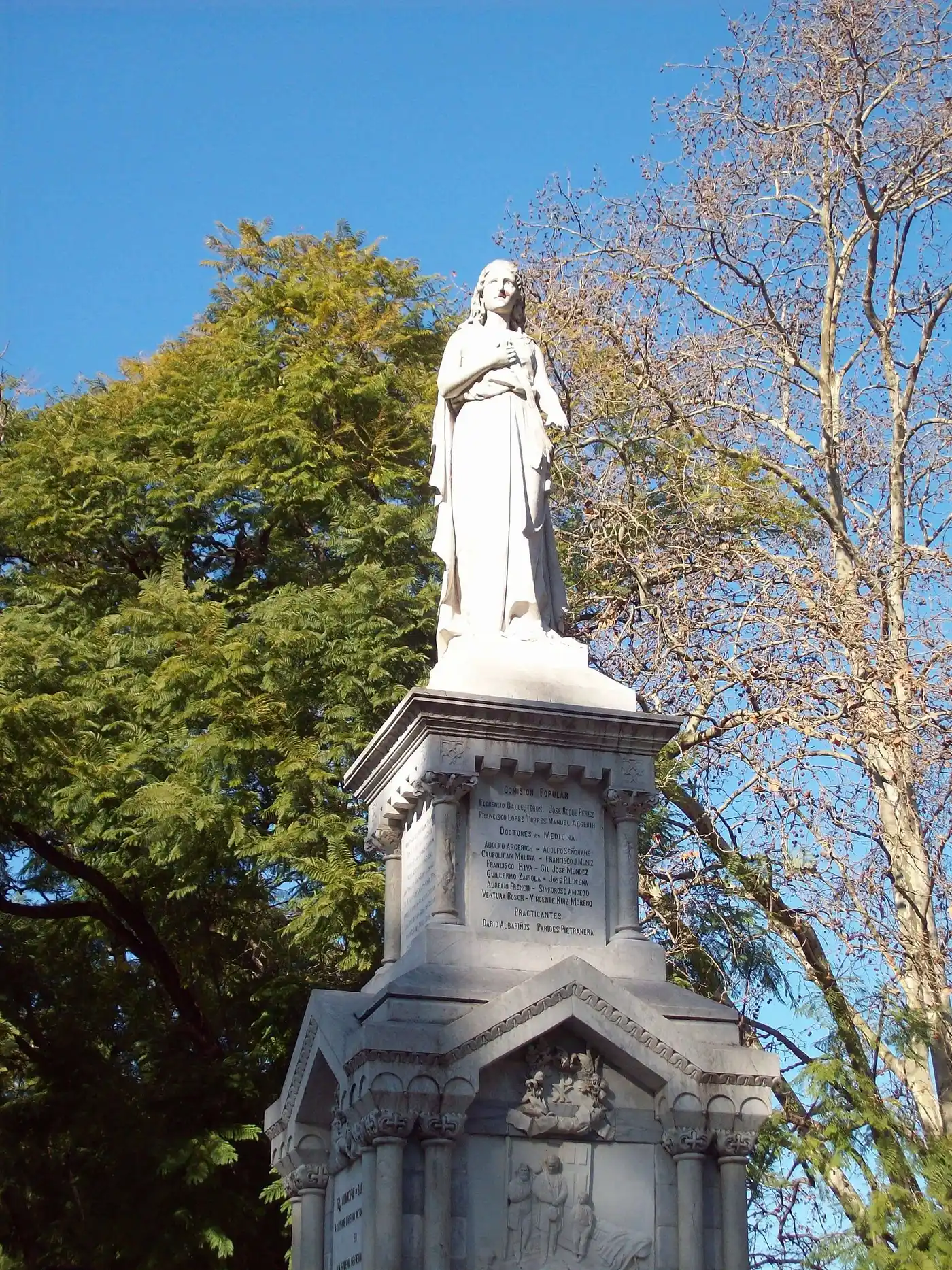 Uma estátua de pedra sobre um pedestal com nomes gravados, entre árvores, sem pessoas.