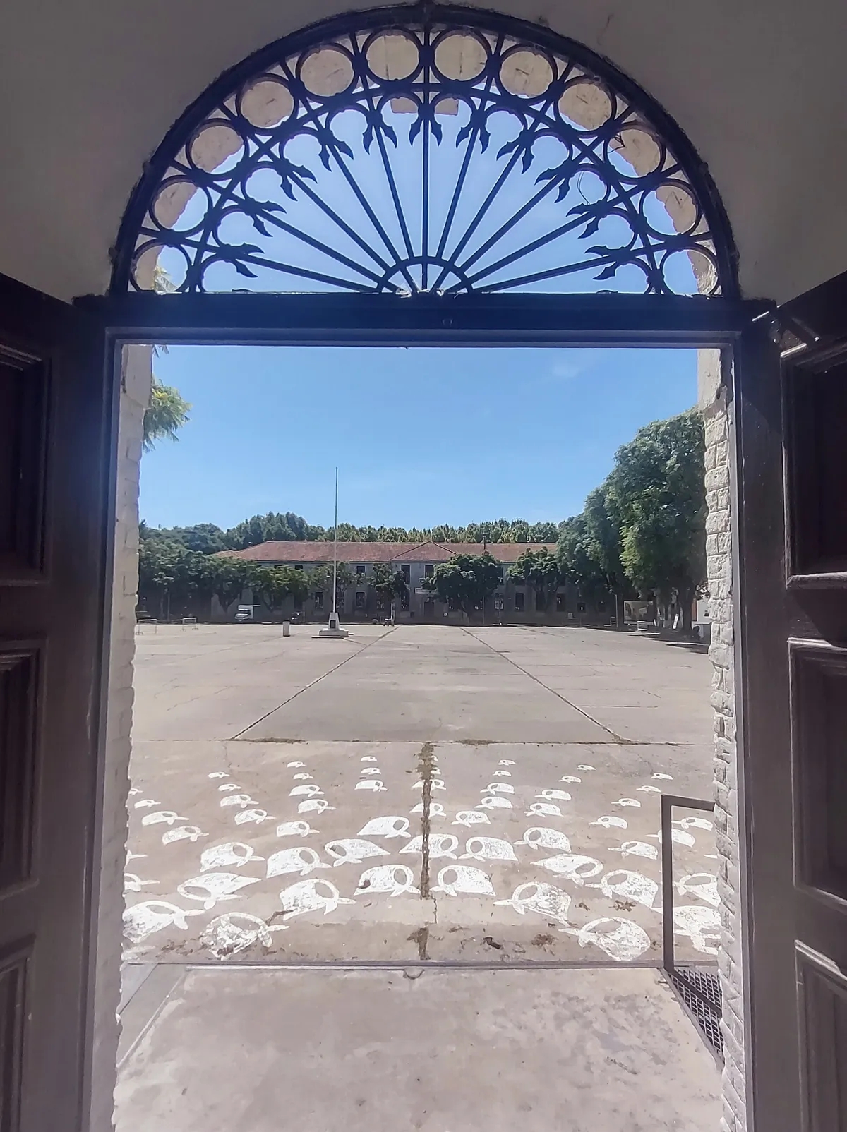 Pátio visto por um leque de ferro, com lenços brancos pintados no chão, um mastro e o telhado longo do edifício ao fundo.