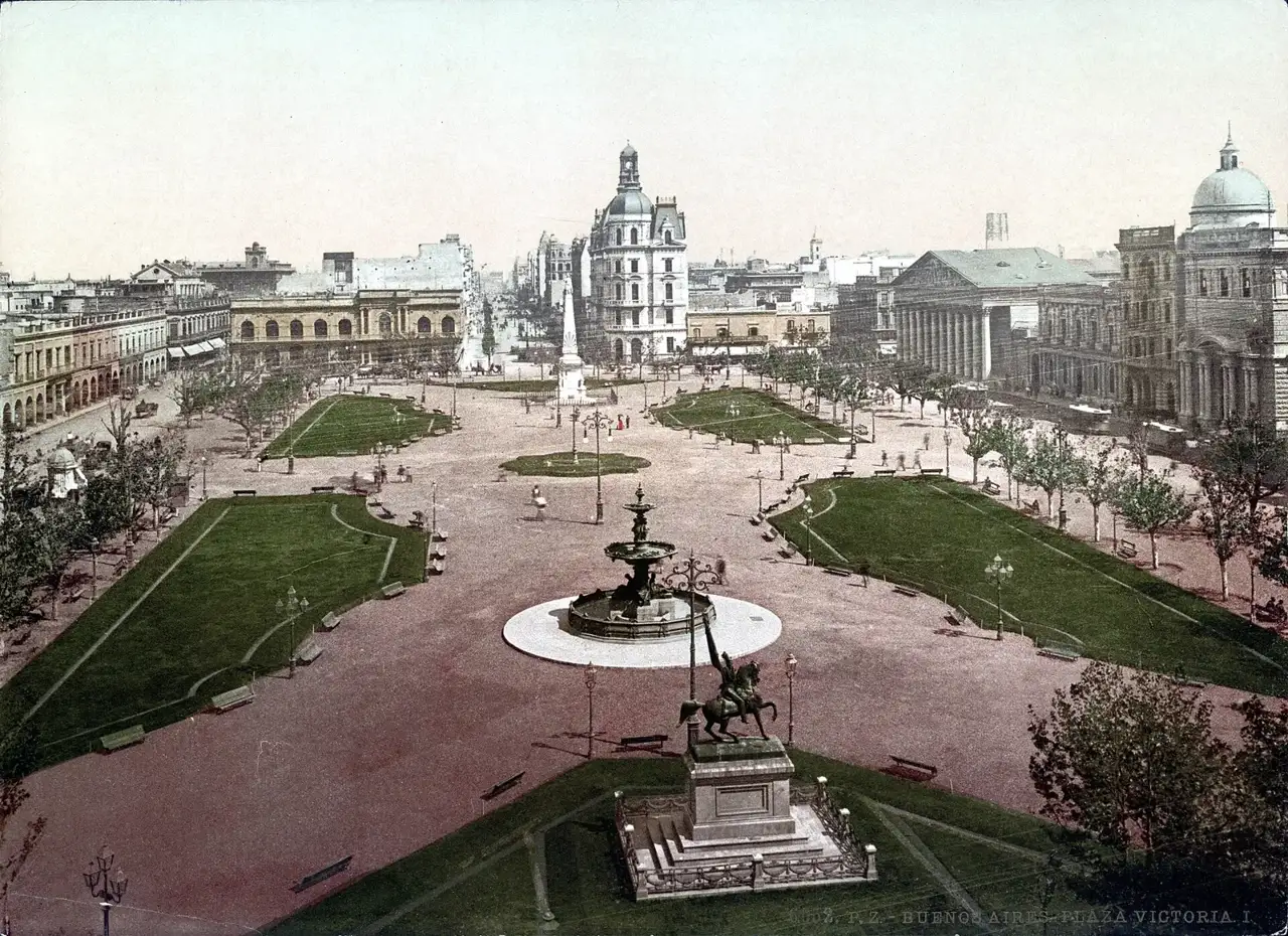 Fotocromo colorido da Plaza Victoria em Buenos Aires, com uma fonte e uma estátua equestre em primeiro plano, gramados, e prédios ao redor da praça.
