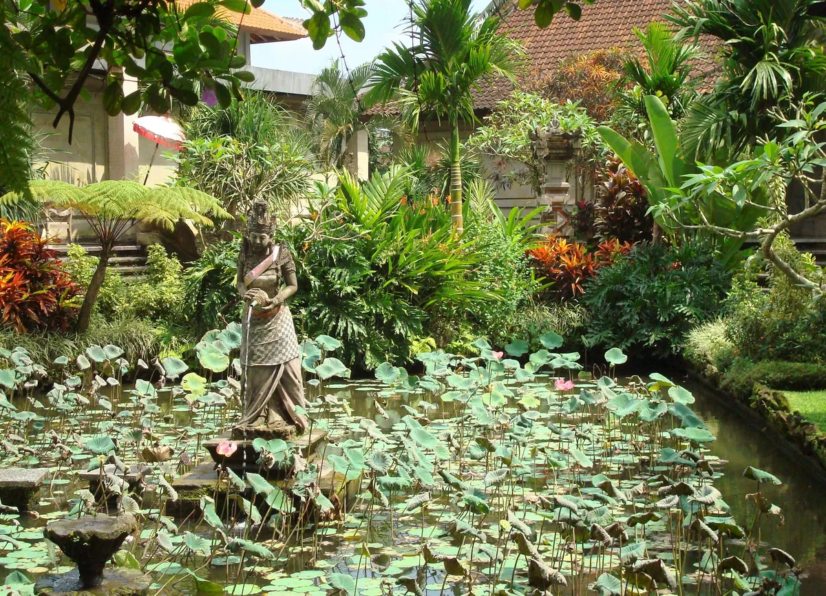 Estátua de pedra no meio de um lago de lótus, com um pavilhão de telhado de telha ao fundo.