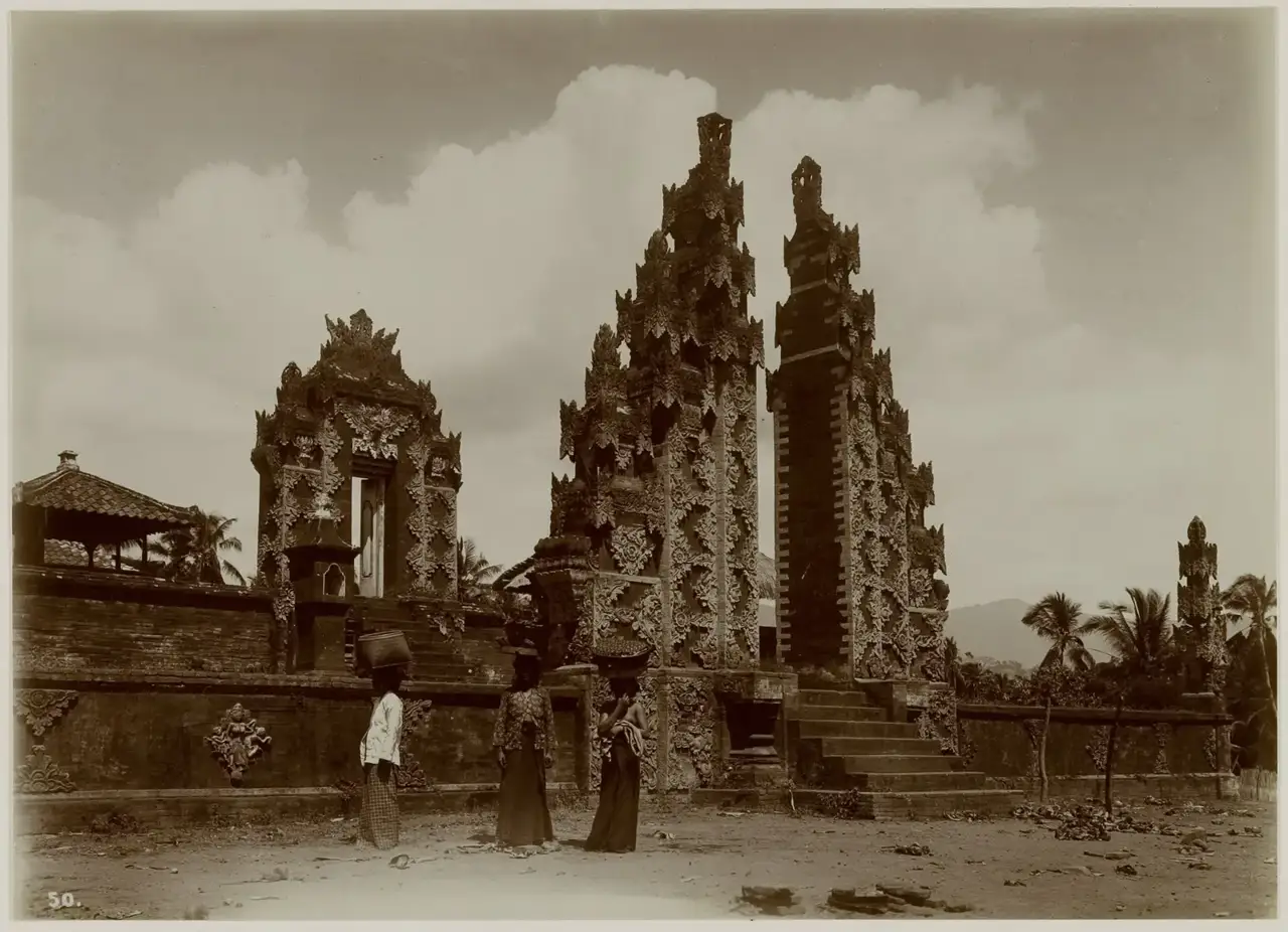 Fotografia em tons sépia de um portal partido (candi bentar) esculpido de um templo em Bali, com três mulheres carregando cestos na cabeça diante da entrada.