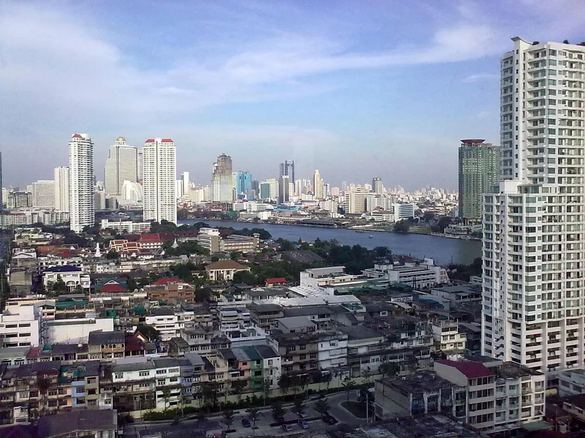 Torres brancas ao longo do Chao Phraya, com o rio no meio e o céu claro.