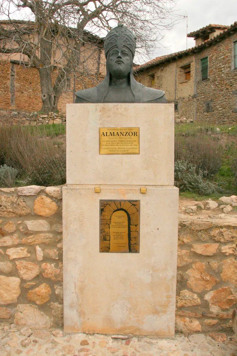 Busto de Almanzor com barba e turbante, sobre pedestal com placas, entre paredes de pedra, vasos e vegetação.