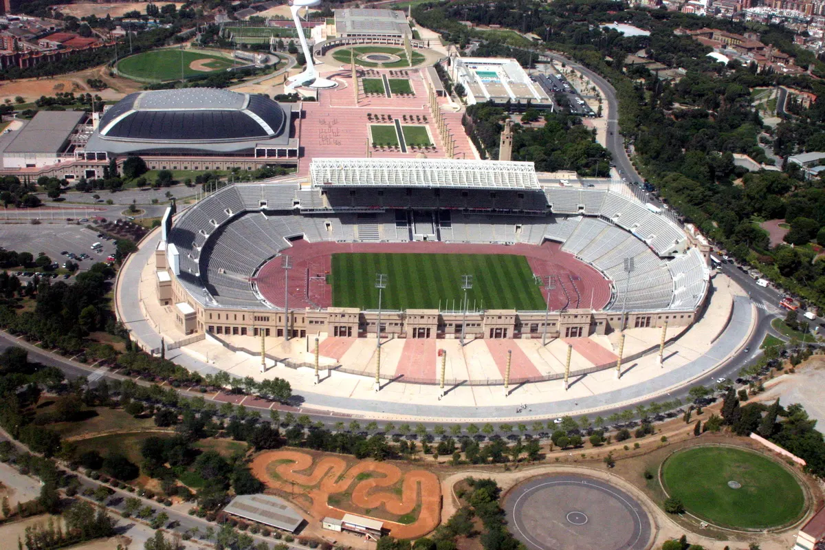 Vista aérea do estádio de Montjuïc, com campo, pista e arquibancadas, diante da esplanada, Palau Sant Jordi e torre de comunicações.