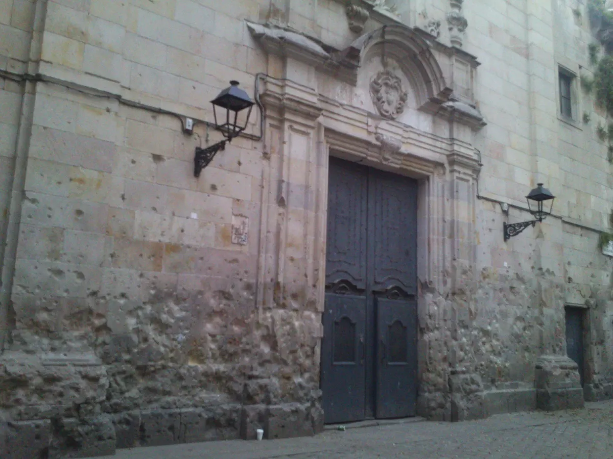 Parede e portal de pedra da igreja de Sant Felip Neri, com numerosas cavidades na alvenaria e calçada diante da fachada.