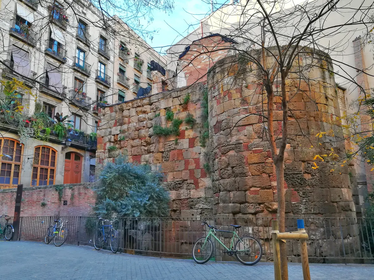 Trecho de muralha e torre circular de pedra na praça Traginers, com árvores, bicicletas e edifícios modernos ao redor.