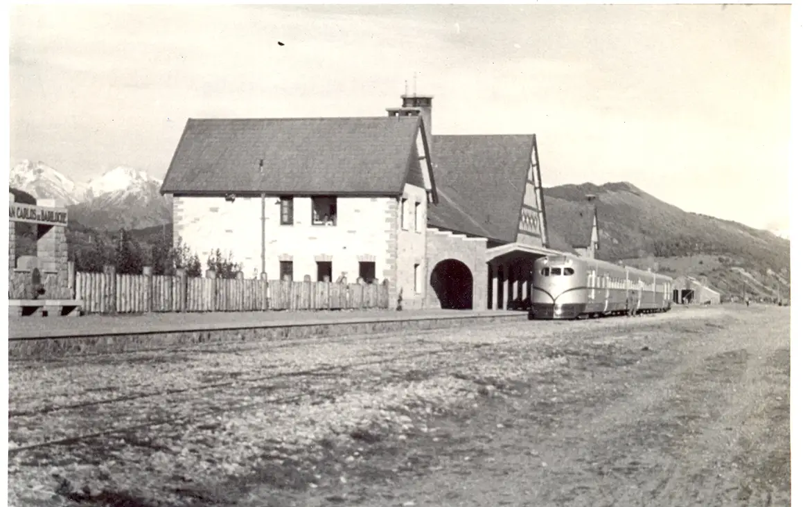 Fotografia em preto e branco da estação de pedra, de telhado íngreme, com um trem claro parado sob a cobertura, uma cerca de madeira e montanhas ao fundo.