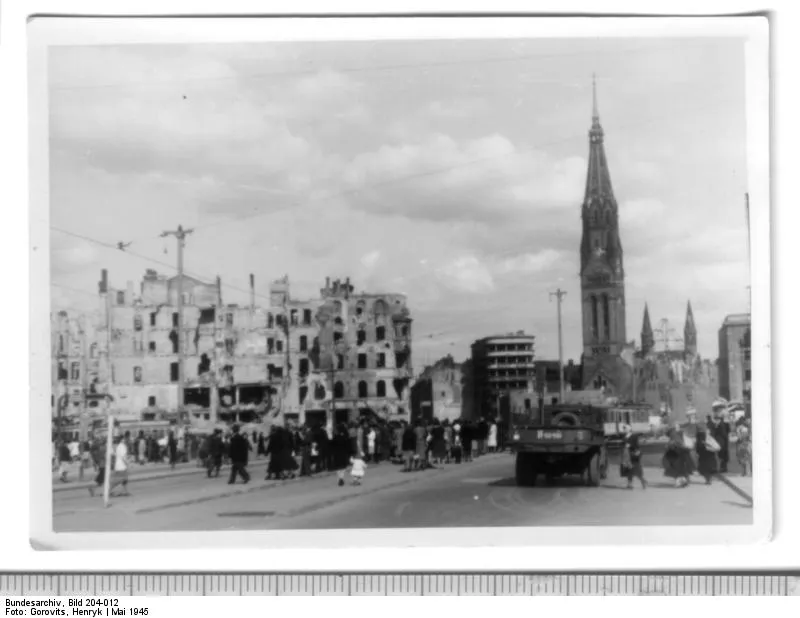Fotografia de edifícios arruinados e da torre da Georgenkirche ao fundo, com bonde, pedestres pequenos e a borda da cópia de arquivo.