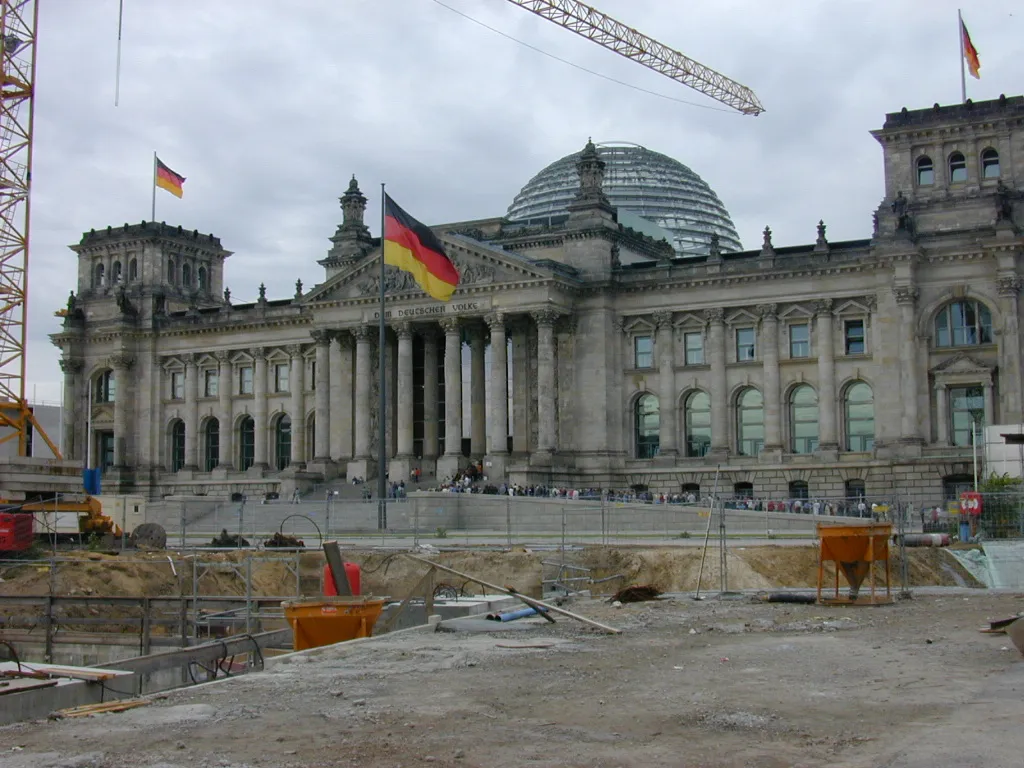 Vista parcial do Reichstag com cúpula de vidro, bandeiras e obras no primeiro plano; visitantes pequenos diante da entrada.