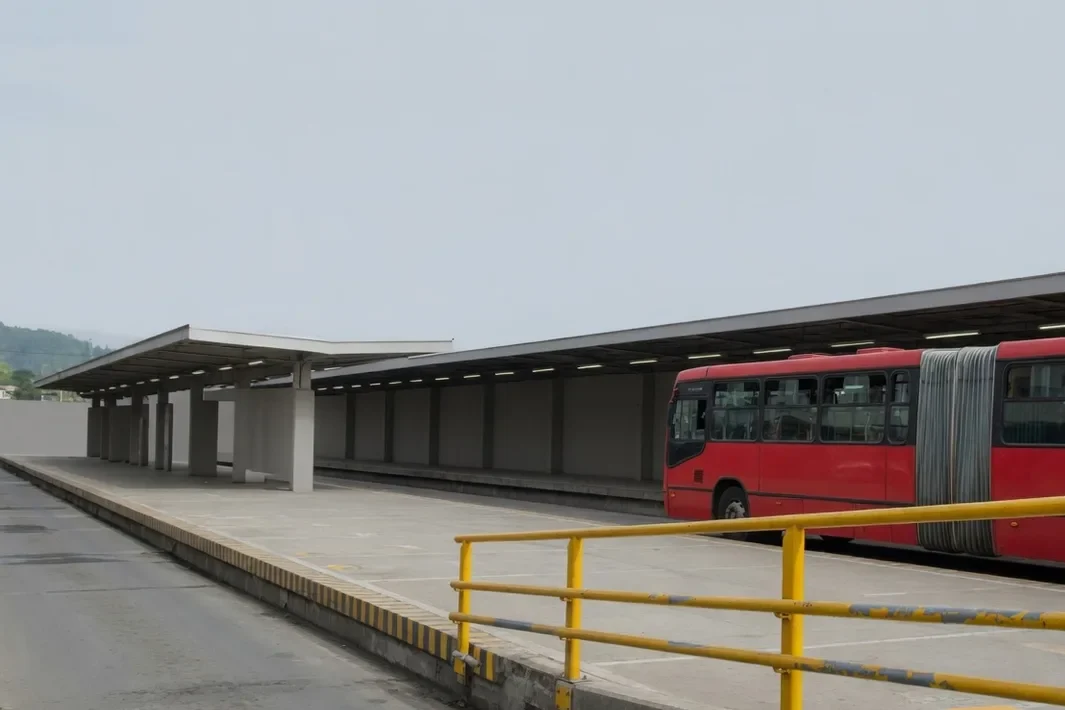 Ônibus articulado vermelho num corredor, com gradil amarelo e céu claro.