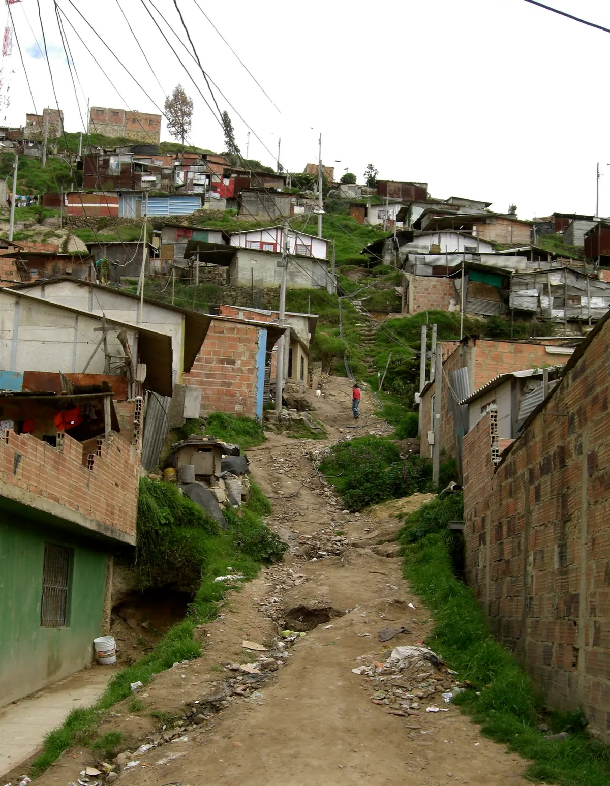 Casas de tijolo e telhas de zinco ao longo de uma trilha de terra na encosta, com uma pessoa de costas ao fundo.