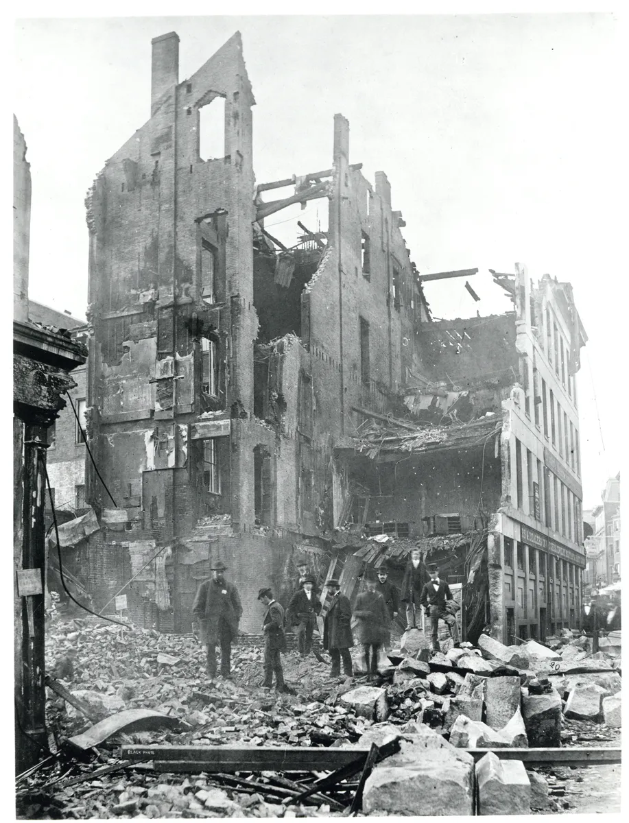 Fotografía de edificios sin tejado, muros de ladrillo, vigas y escombros, con hombres entre los restos y bordes claros.