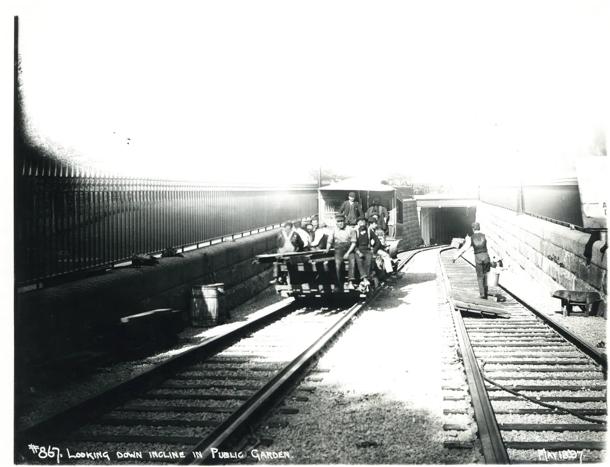 Fotografía de trabajadores sobre un vagón ante la entrada del túnel, vías, rejas, tablas e inscripción fechada en el margen inferior.