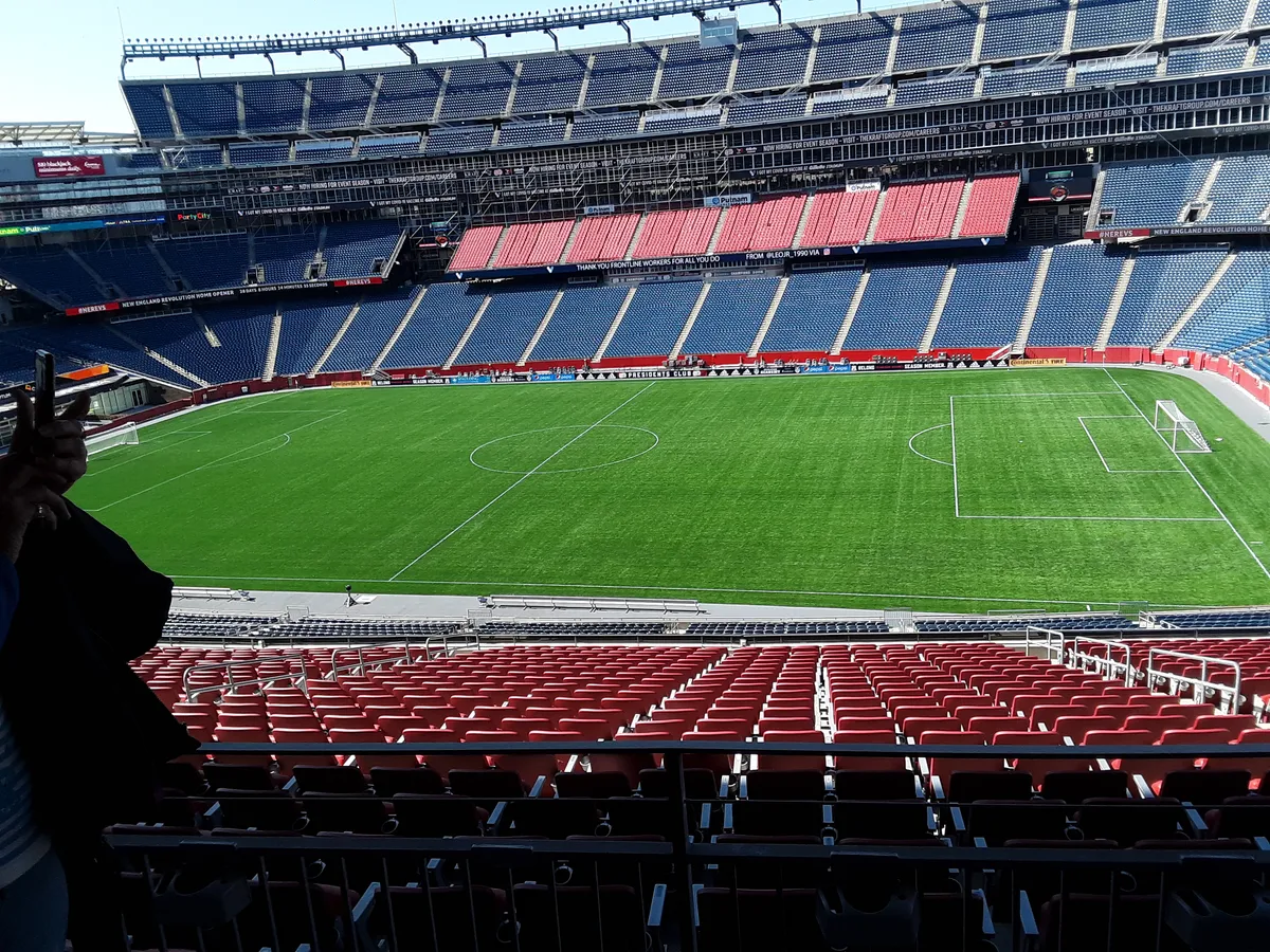 Vista das arquibancadas vermelhas e azuis e do campo vazio do estádio, com traves e mão segurando um telefone no canto esquerdo.