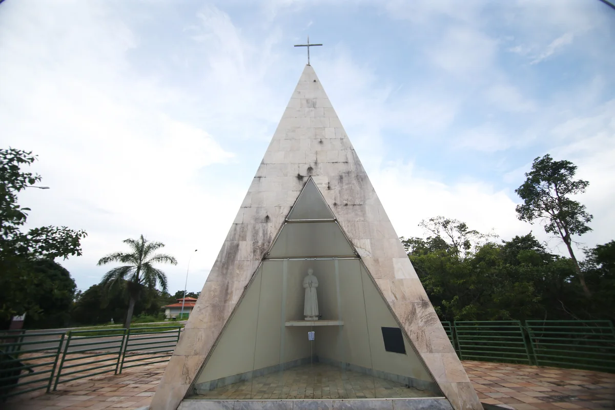 Ermida de pedra em forma triangular, com cruz no alto, escultura no nicho, piso e árvores ao redor.