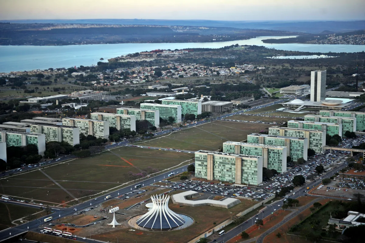 Vista aérea da Esplanada dos Ministérios, com catedral no primeiro plano, edifícios ministeriais alinhados, Congresso e lago ao fundo.