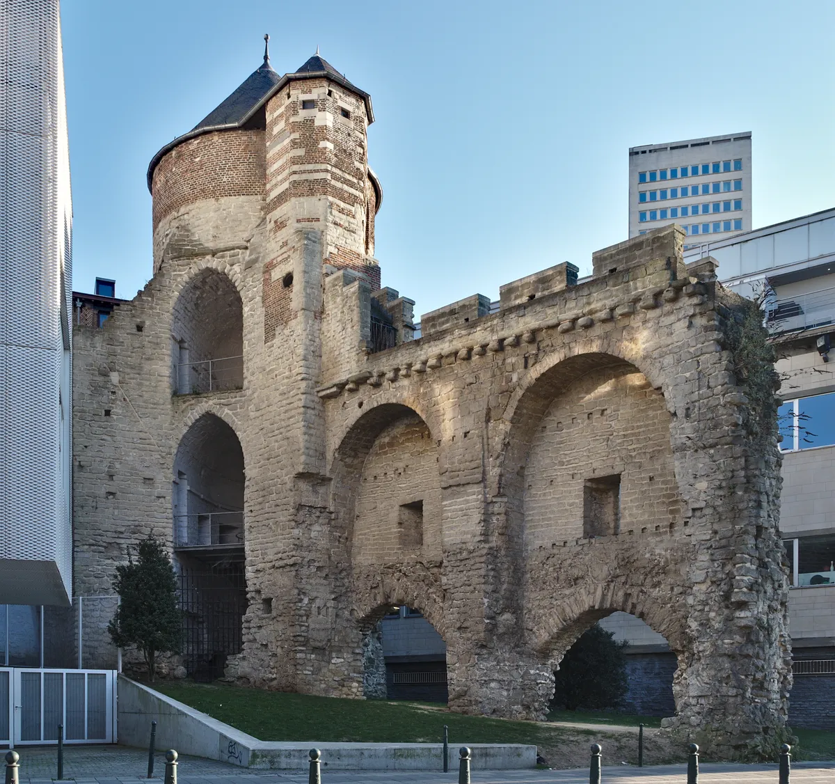 Torre de pedra e tijolo com telhados pontudos, arcos da muralha e edifícios modernos ao redor, vistos da rua.