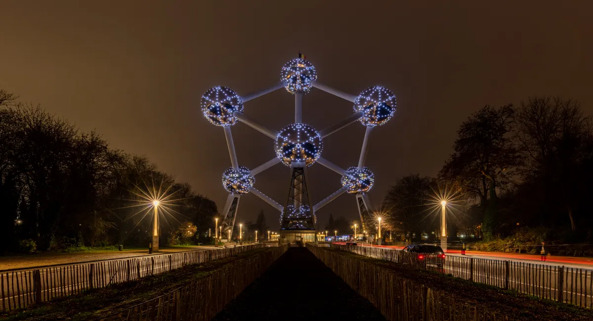 Atomium iluminado à noite, com esferas e tubos metálicos, base, árvores, cercas e rastros de luz dos veículos na avenida.