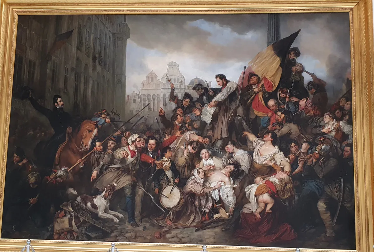 Pintura de uma multidão diante do edifício gótico, com documento, bandeira belga, armas e figuras feridas, dentro da moldura dourada.