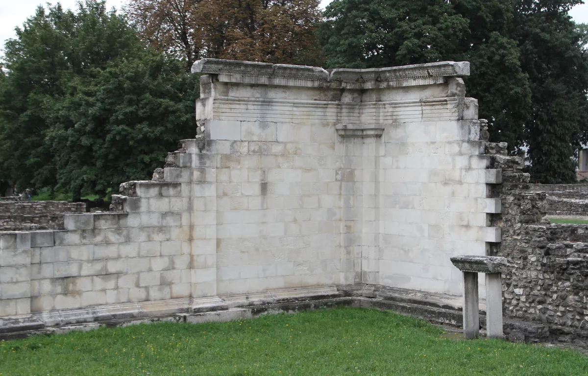 Trecho de paredes romanas de pedra em Aquincum, com uma abertura, gramado e árvores ao fundo.