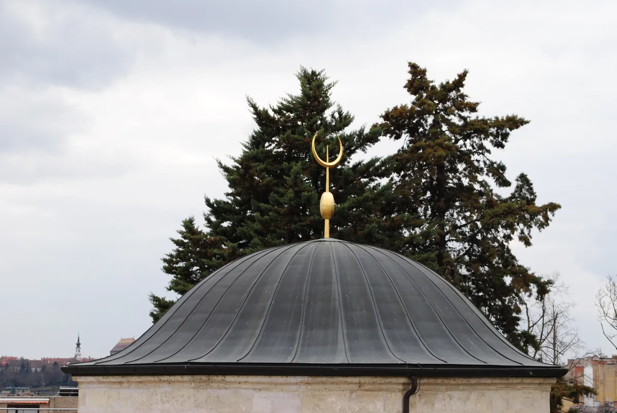 Detalhe da cúpula revestida de metal e do crescente dourado do túmulo de Gül Baba, com árvores e parte da cidade ao fundo.