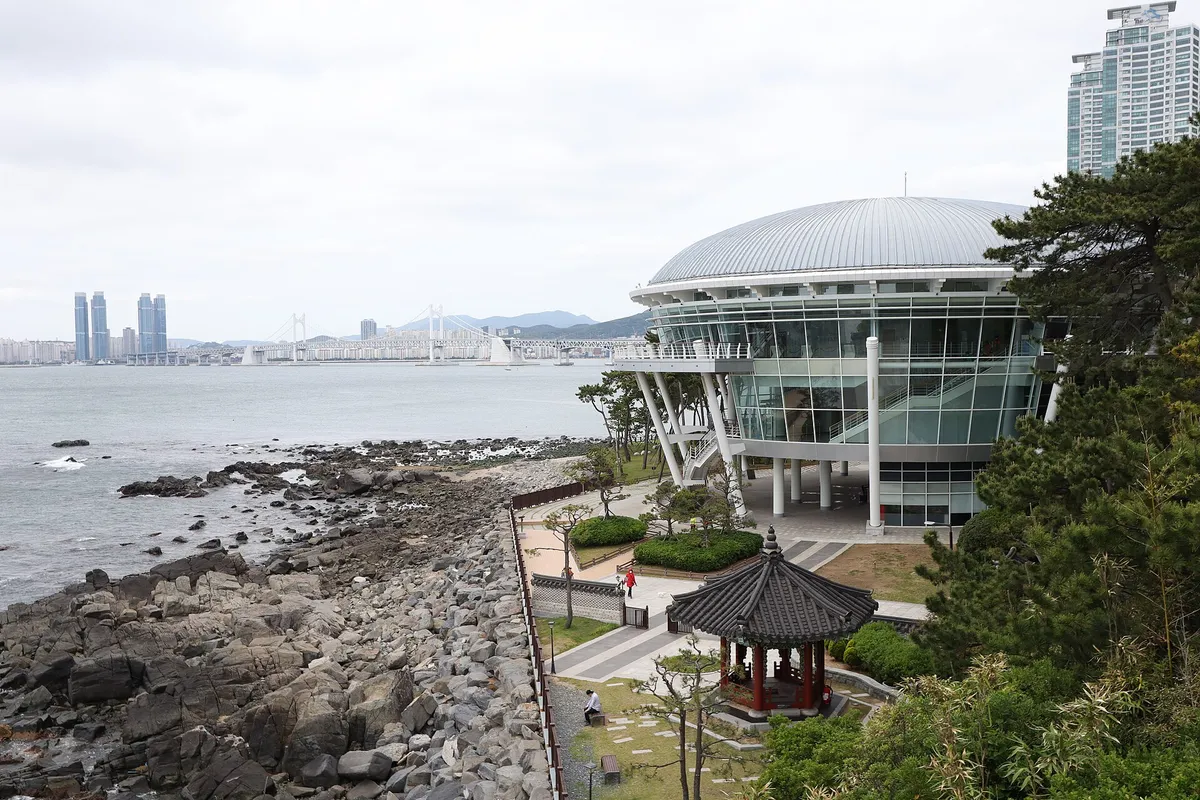 Um edifício redondo de vidro e cúpula metálica, à beira da água, com um pavilhão de telhado curvo na frente.