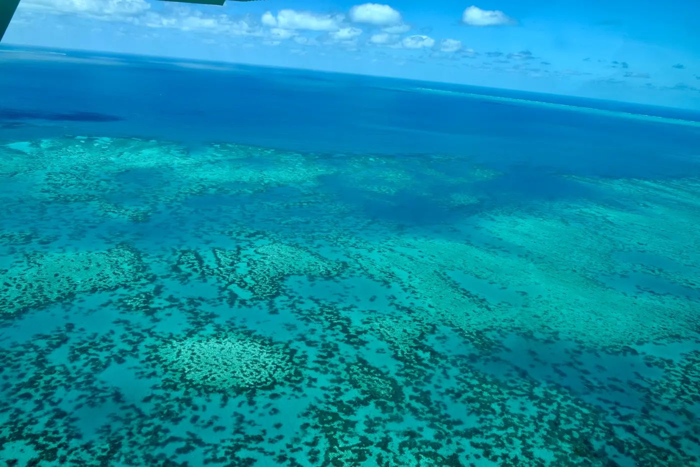 Vista aérea de recifes turquesa sob o mar azul, com nuvens e a borda de uma janela no canto.