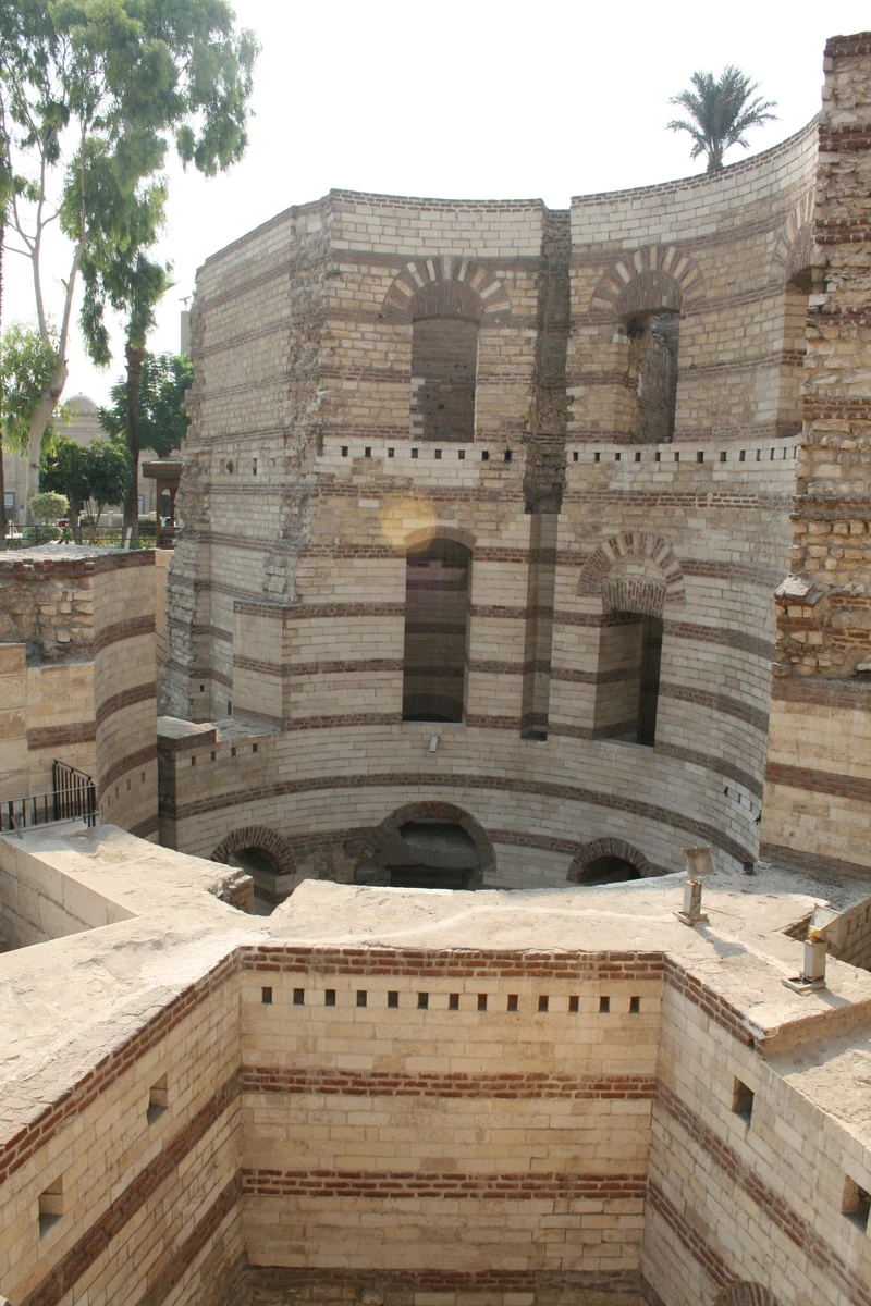 Torre curva de pedra e tijolo, com aberturas em arco e ruínas em primeiro plano.