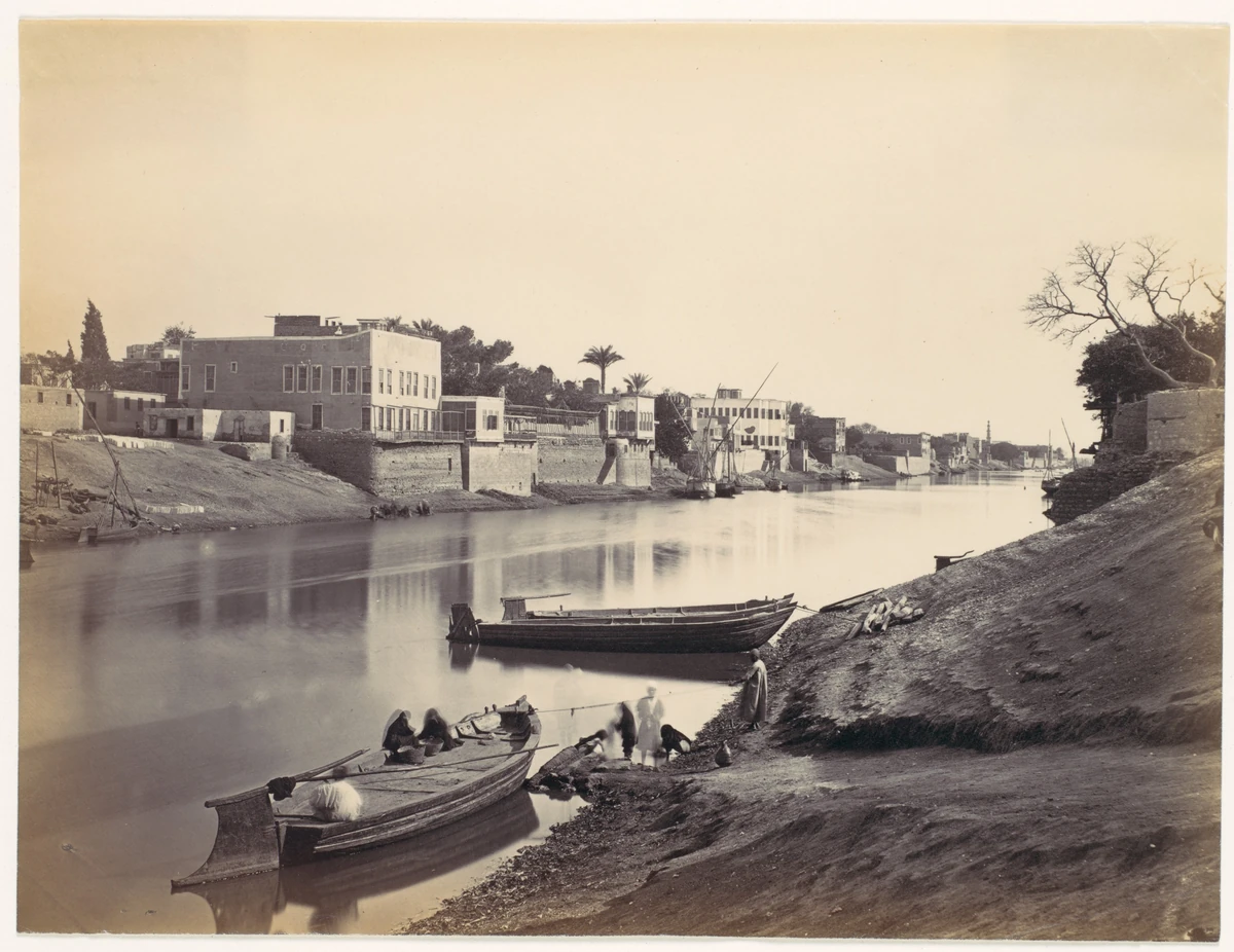 Fotografia sépia do rio entre margens com edifícios, barcos junto à margem e pessoas em primeiro plano.