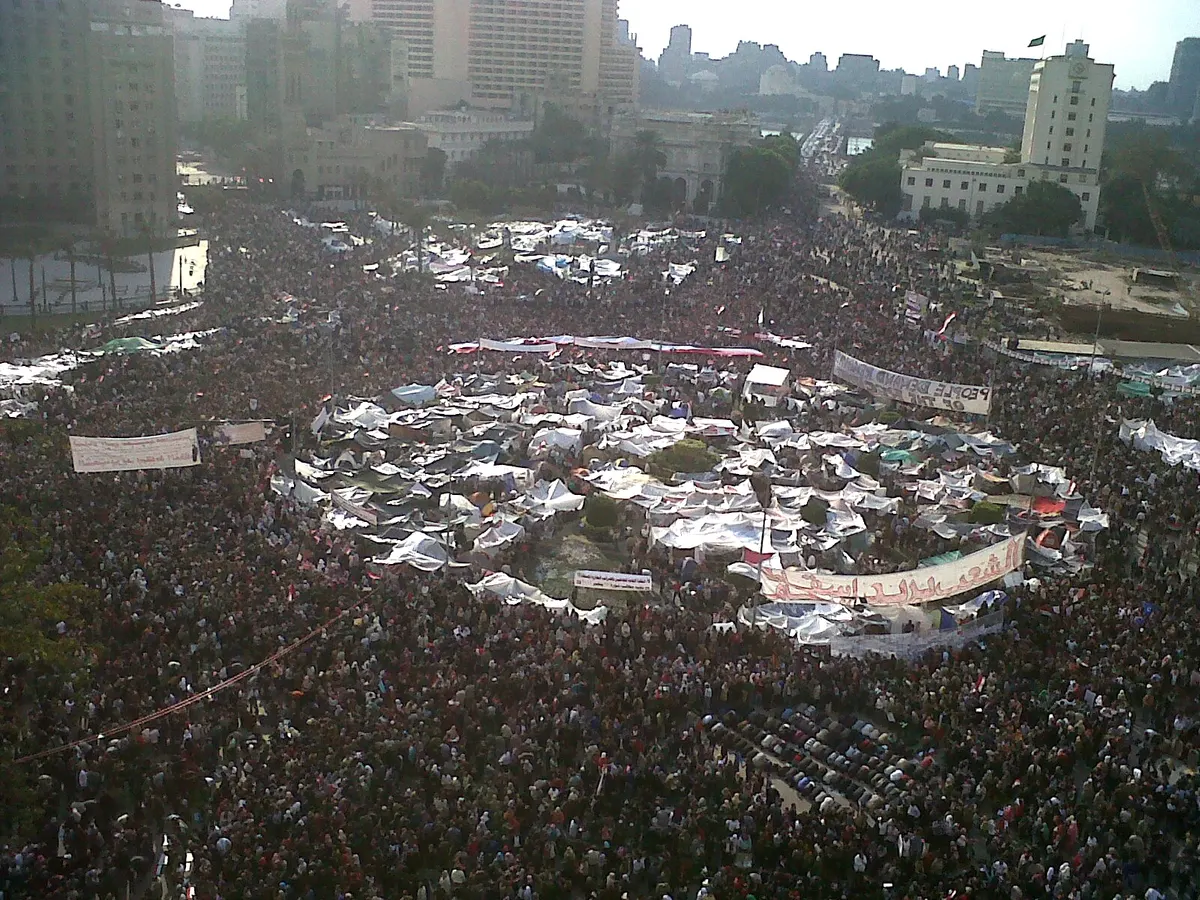 Vista elevada da Praça Tahrir cheia de manifestantes, tendas e faixas, com árvores e edifícios ao fundo.