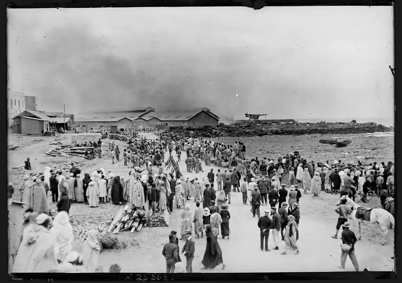 Fotografia em preto e branco mostra uma multidão observando a fumaça de um incêndio perto dos armazéns do porto de Casablanca, com soldados europeus e marroquinos de djellaba nas ruas.