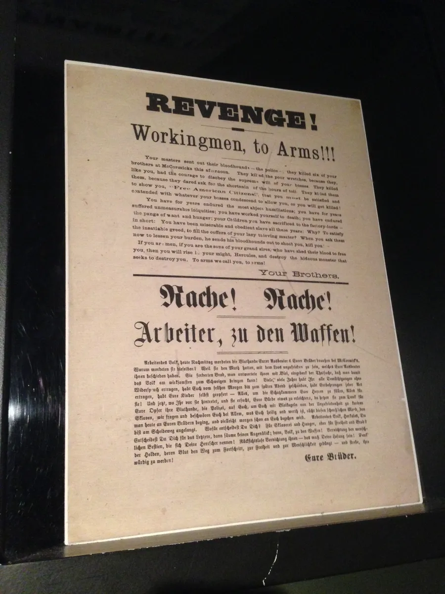 Photographie d’une feuille imprimée exposée, avec titres en anglais et allemand, texte dans les deux langues et reflets sur fond sombre.