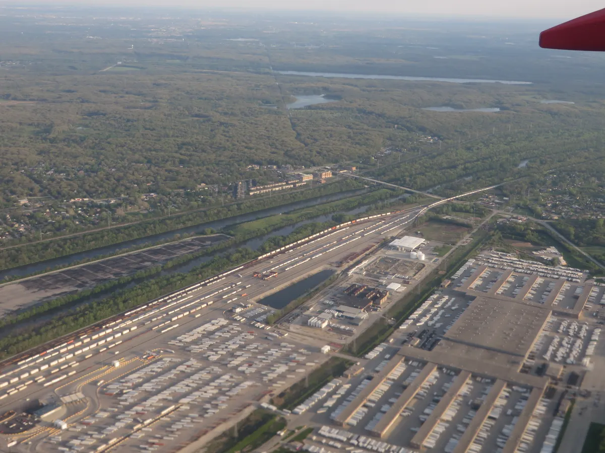 Vista aérea de canal entre faixa de árvores, linhas ferroviárias, galpões e ruas, com parte da asa do avião no canto superior direito.
