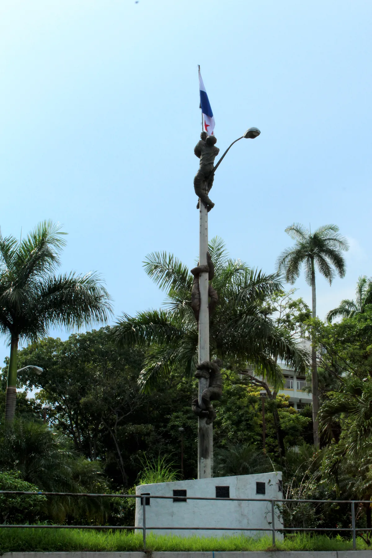 Três figuras de bronze sobem um mastro com a bandeira do Panamá, sobre uma base branca, entre palmeiras.
