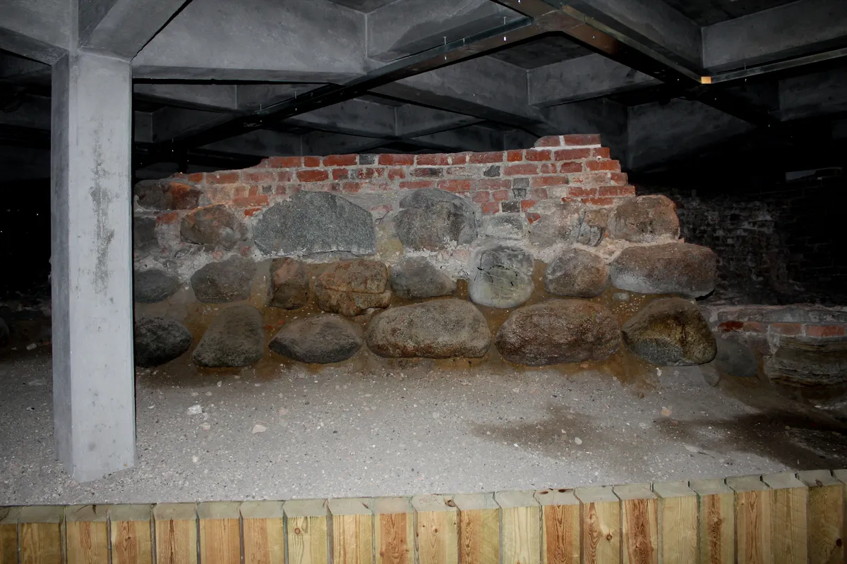 Ruínas com pedras arredondadas e tijolos vermelhos, sob vigas modernas, com coluna à esquerda e barreira de madeira na frente.