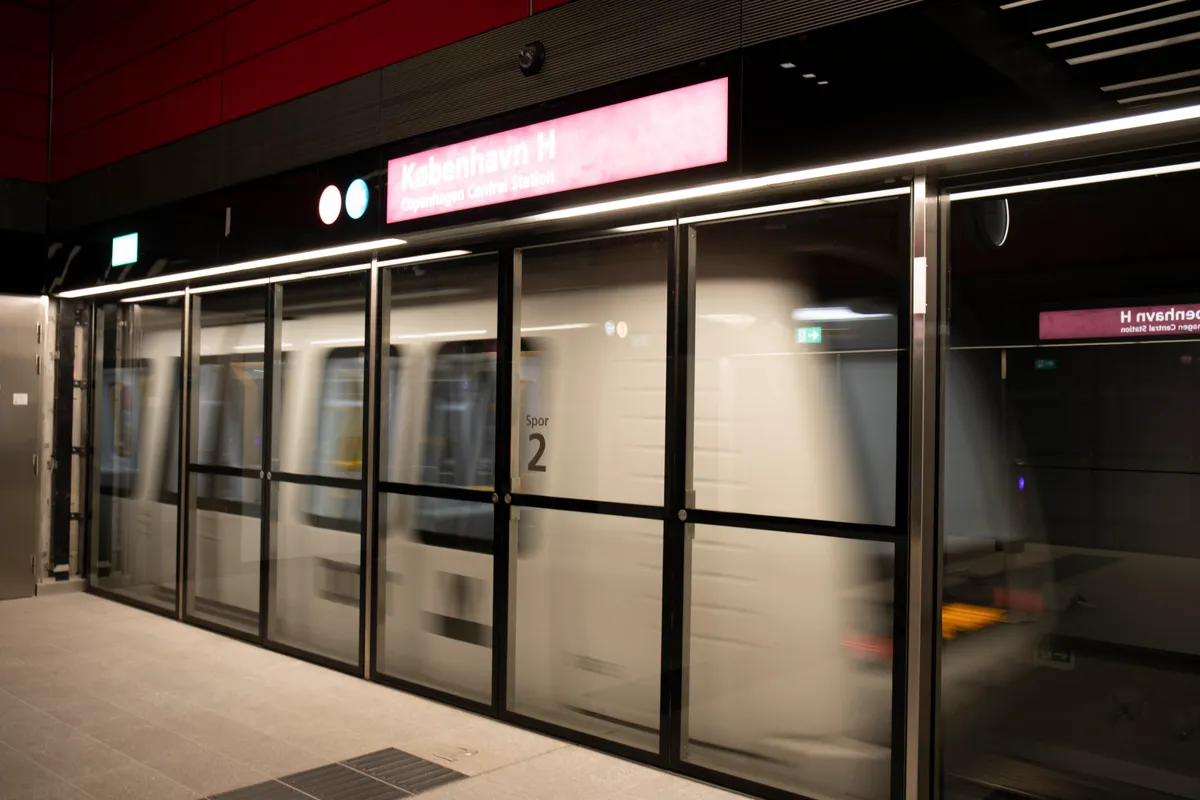 Estação com o nome København H, portas de vidro junto à plataforma, trem ao fundo, faixa amarela no piso e sinal indicando a via 2.