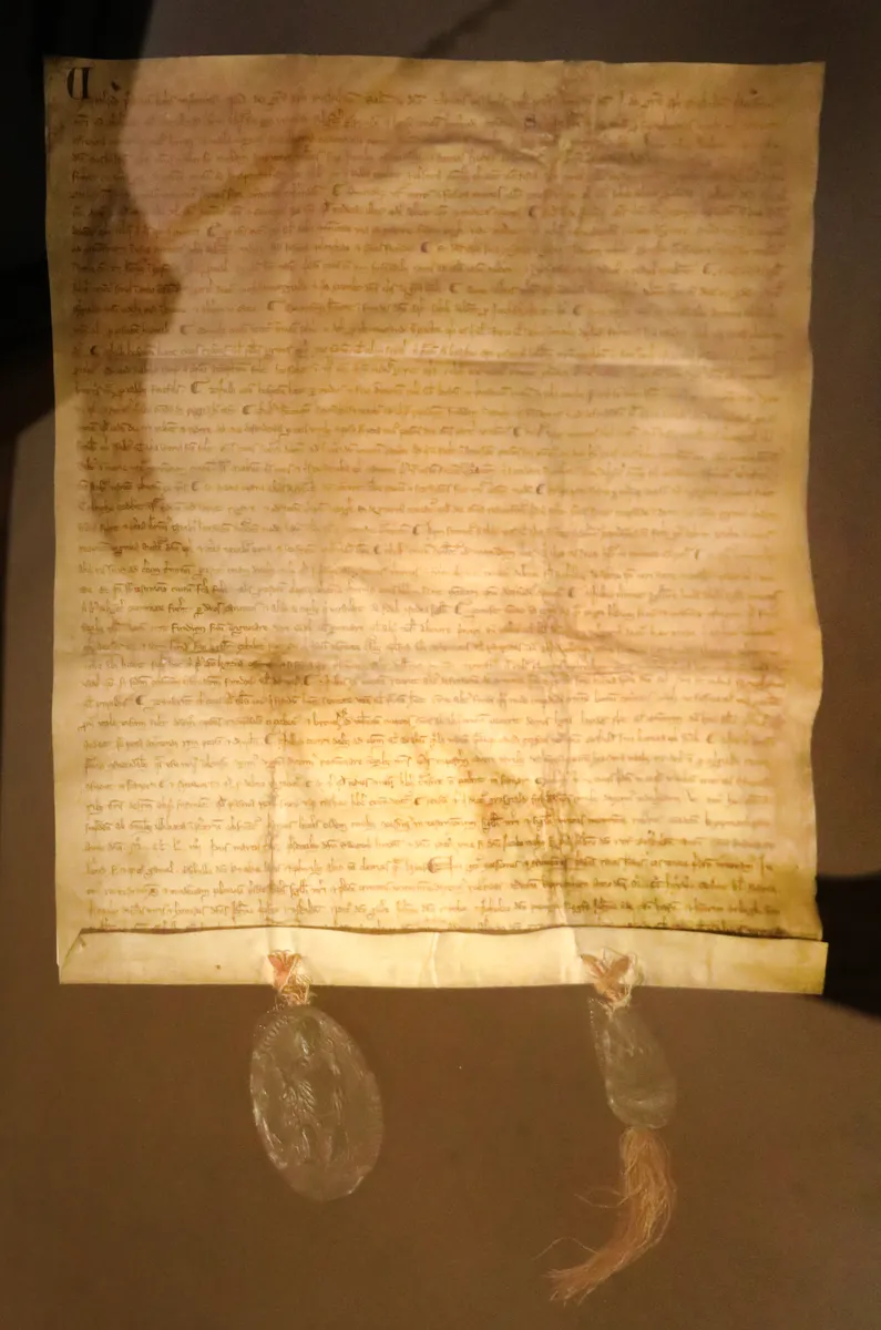 Documento manuscrito em pergaminho com duas faixas suspensas e selos parcialmente quebrados, sobre fundo cinza, com reflexos do vidro.