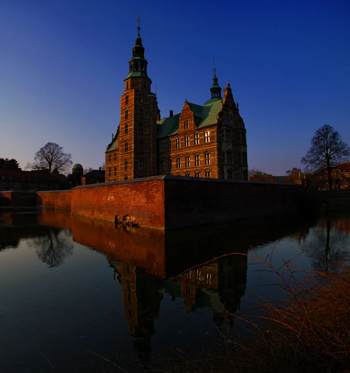 Castelo de tijolos iluminado pelo sol, com torres e telhados completos, fosso refletindo a fachada e ramos no primeiro plano.