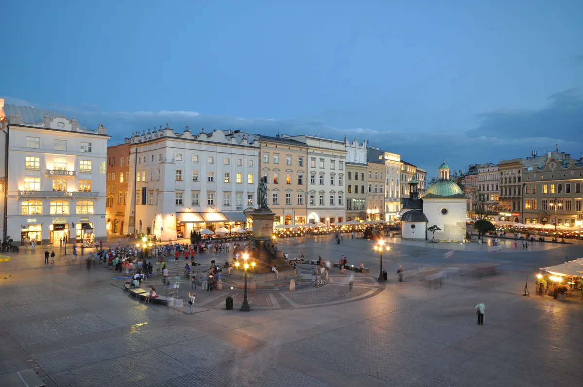 Praça principal ao entardecer, com fachadas iluminadas, monumento de Adam Mickiewicz, pequena igreja com cúpula e pessoas em longa exposição.