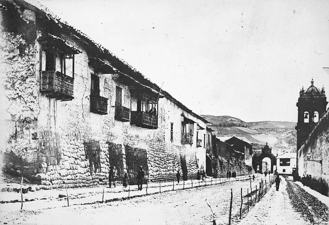 Fotografia em preto e branco mostra uma rua estreita de Cusco com muros de pedra inca sustentando andares coloniais de adobe e sacadas de madeira, e uma torre de igreja ao fundo.