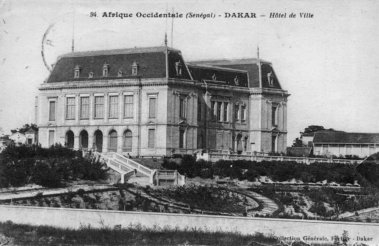 Cartão-postal em preto e branco mostra o Hôtel de Ville de Dakar, um edifício colonial com telhado de mansarda e janelas em arco no térreo, com um jardim e uma escadaria em frente.