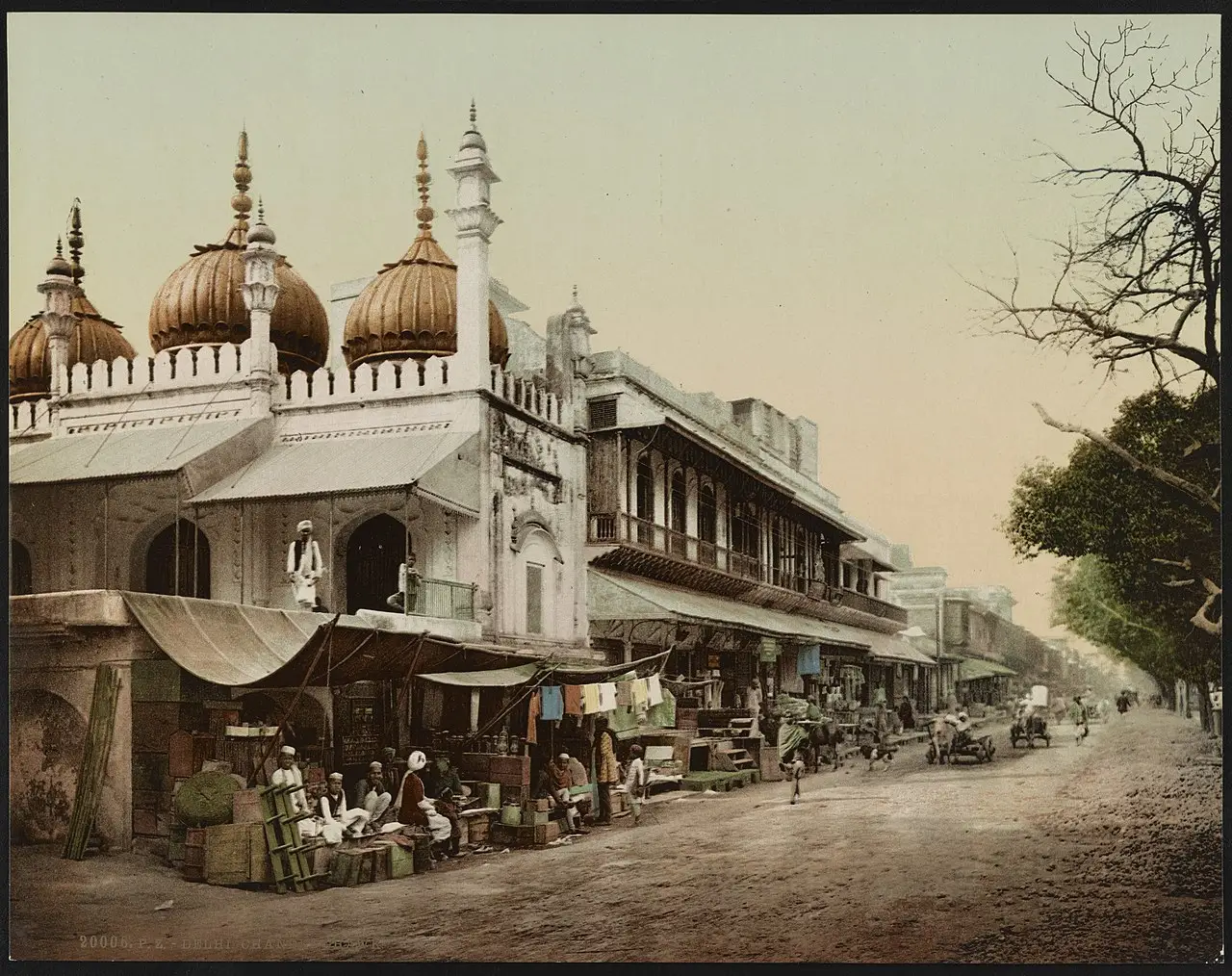 Fotocromo de uma avenida larga: à esquerda um prédio branco com cúpulas e um minarete, embaixo lojas com toldos, gente e carroças, e árvores à direita.