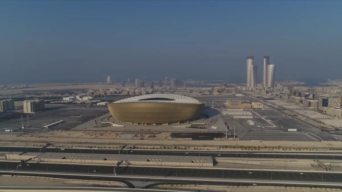 Vista aérea do estádio dourado em forma de tigela, com torres altas ao fundo e a autoestrada em primeiro plano.