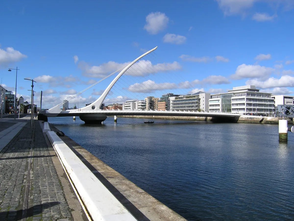 Ponte branca sobre o Liffey, com mastro curvo completo, cabos, tabuleiro, margem do rio e prédios ao fundo.