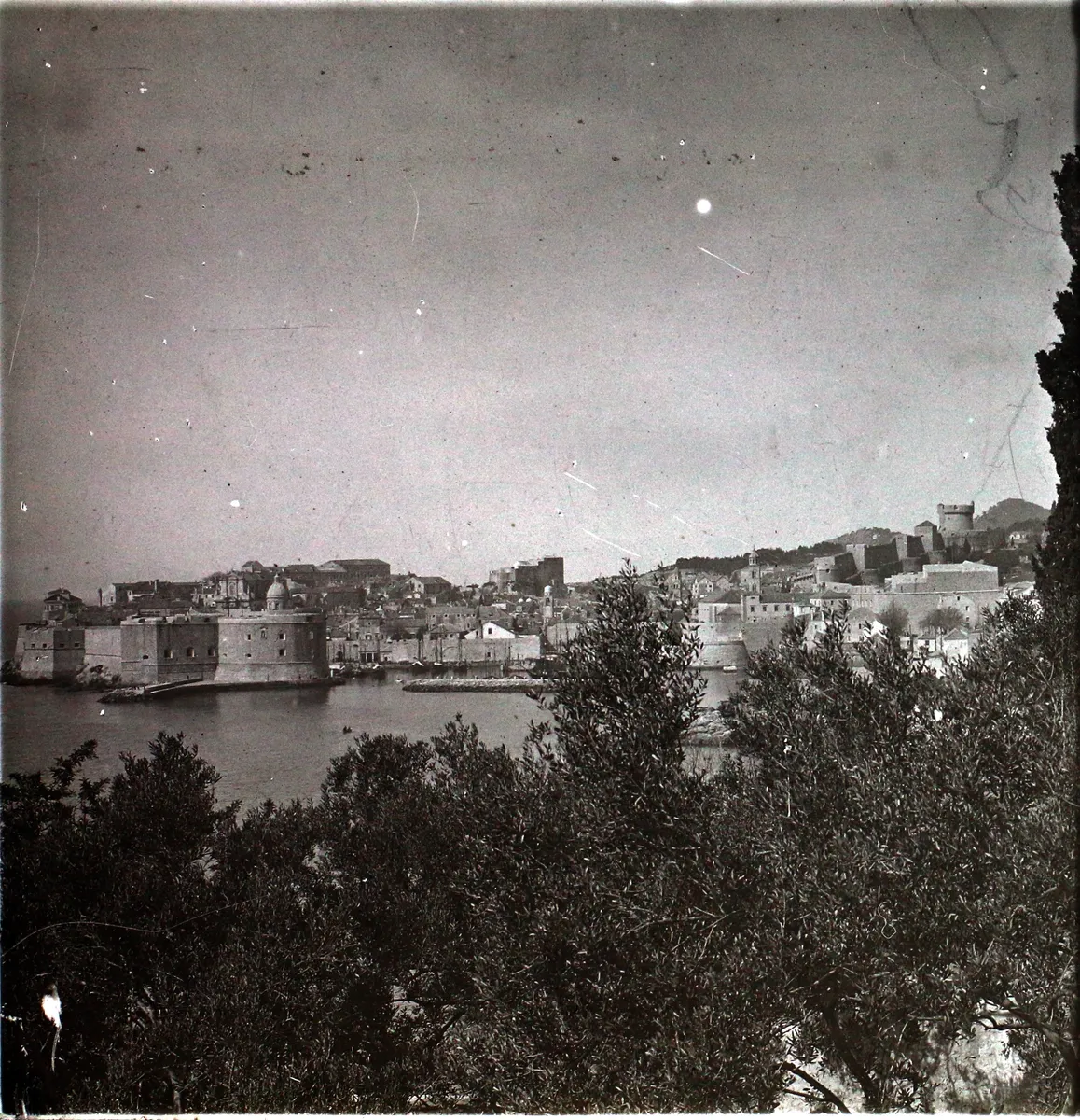 Fotografia vertical com ampla área de céu, cidade murada junto ao mar na parte inferior, telhados, embarcações e galhos em primeiro plano, com bordas e marcas do suporte.
