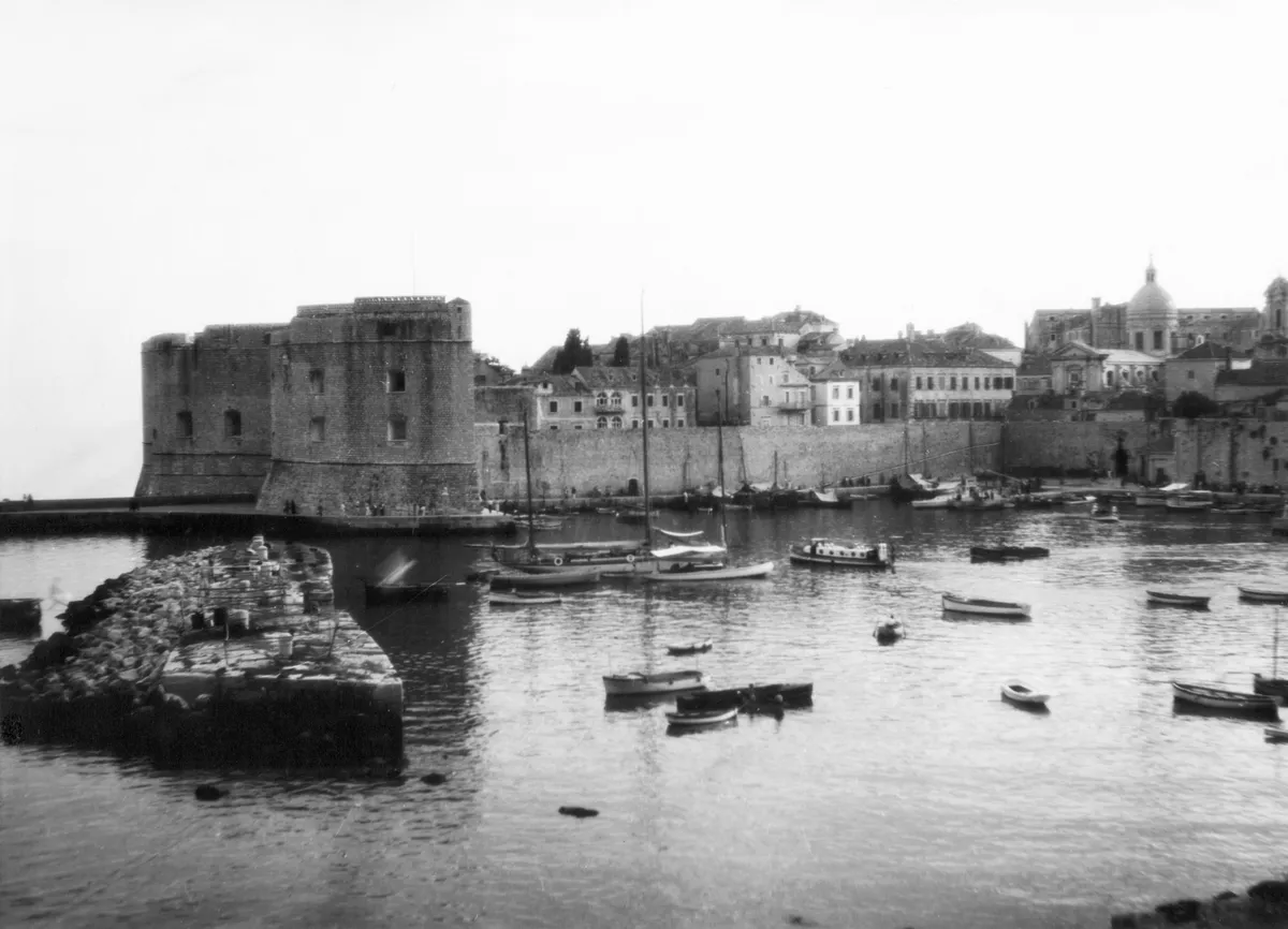 Fotografia do porto com muralhas, telhados e cúpula ao fundo, embarcações na água, quebra-mar de pedra e pequenas figuras junto à costa.