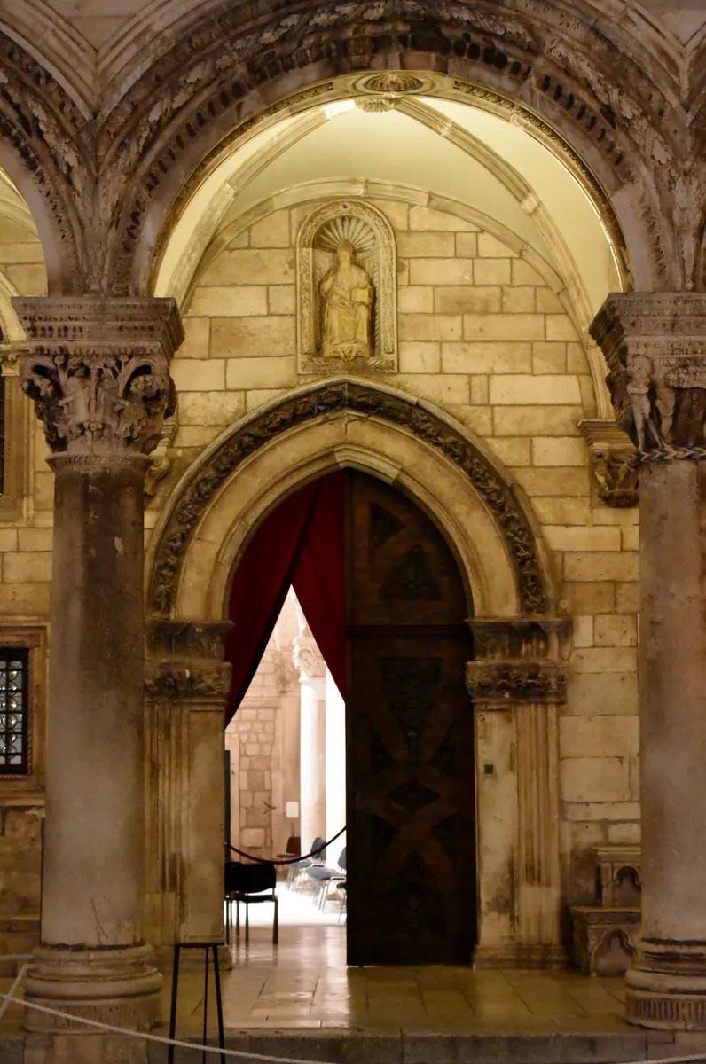 Portal de pedra iluminado à noite, arco e colunas com capitéis, figura esculpida no nicho, porta entreaberta, cortina vermelha e cordão à frente.
