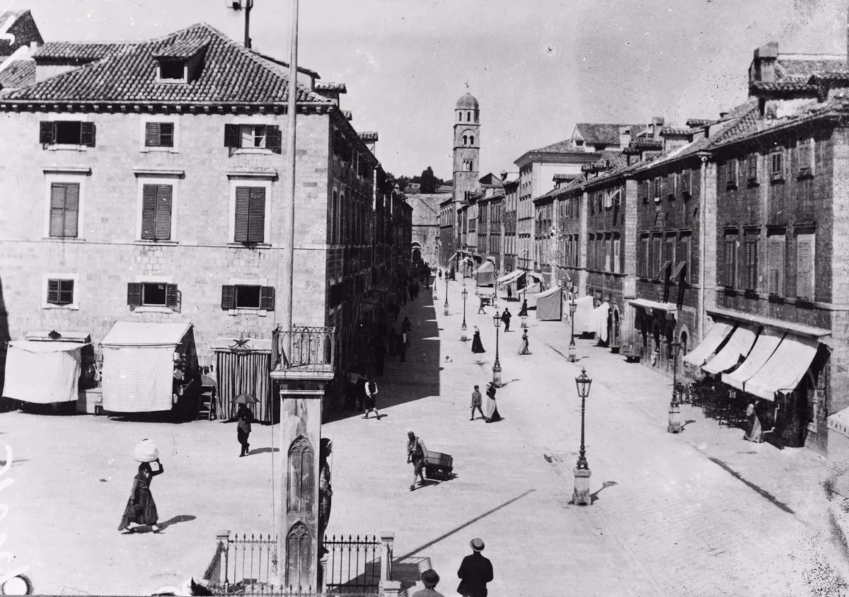 Fotografia do Stradun com rua pavimentada, fachadas, campanário ao fundo, coluna esculpida, toldos, postes e pequenas figuras, dentro das bordas da imagem histórica.