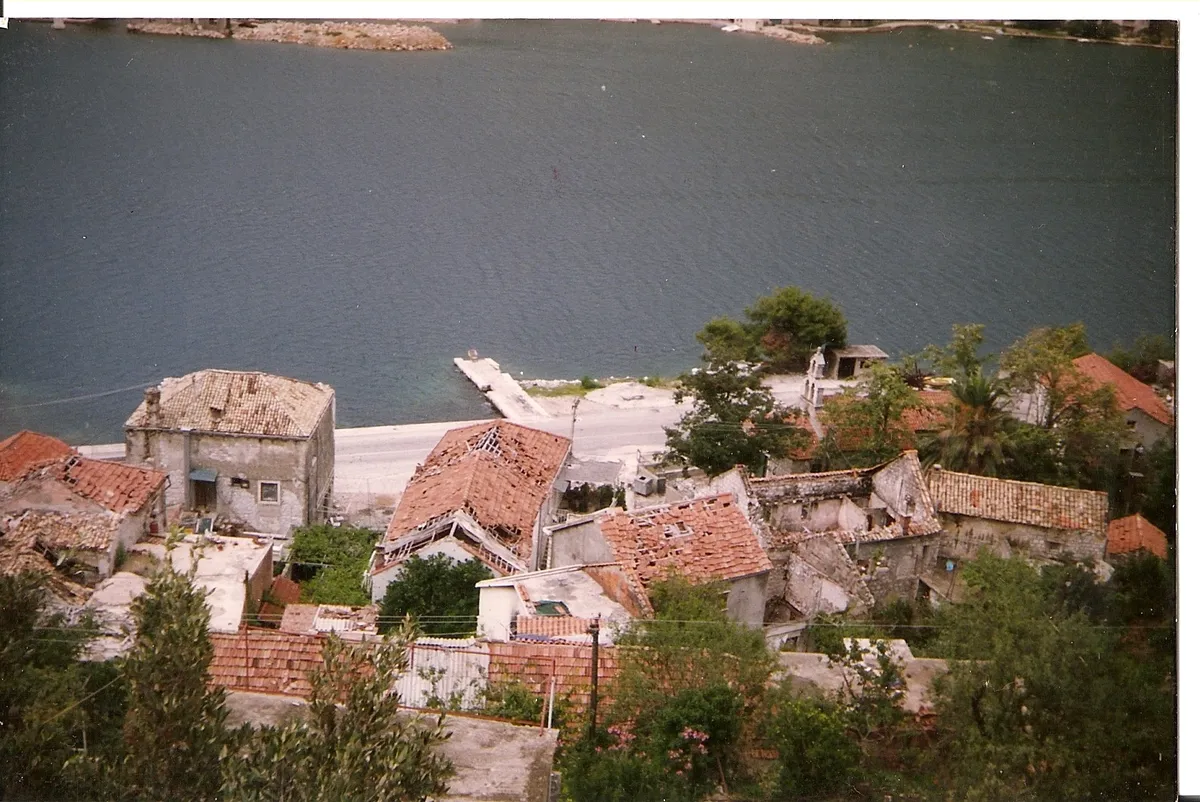 Fotografia de telhados danificados com vigas expostas e paredes de pedra, árvores, via costeira e água ao fundo, rodeada pelas margens claras da cópia.
